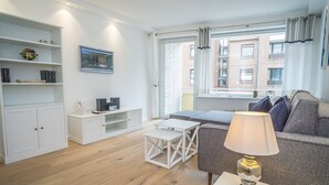 Un soggiorno moderno con un divano grigio, un tavolino da caffè bianco e una lampada. Si nota un mobile bianco con scaffali e una televisione su un supporto. Si vede una finestra con tende e un edificio in mattoni all'esterno.