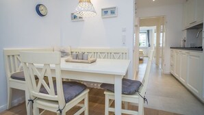 Zona pranzo con tavolo bianco, due sedie, una panca, un lampadario decorativo e un orologio appeso al muro.