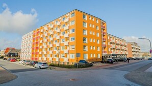 A multi-story hotel with a distinctive orange facade, multiple balconies, and a parking area in front. - Windhuk 1-11 am Strand (Westerland)
