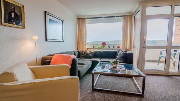 A living room with a sofa, armchair, and coffee table. There is a framed picture on the wall and a window with curtains. - Windhuk 1-11 am Strand (Westerland)