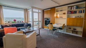 A modern living room with a sofa, coffee table, and dining area. There is a bookshelf with books and decorative items. A large window provides a view of the outdoors. - Windhuk 1-11 am Strand (Westerland)