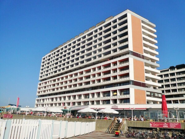 A multi-story hotel with a grid-like facade, balconies on each floor, and a covered outdoor seating area with umbrellas. - Haus Metropol, App. 276a OB Lage: Das Haus Metropol Steht Direkt am Strand von Westerland Zwischen den Beiden Fußgängerzonen. In Unmittelbarer Nähe Befinden Sich die "sylter Welle" und das "syltness-center". Das Appartement Befindet Sich in der (Westerland)