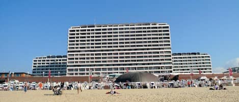 A large hotel with multiple floors and balconies, situated on a sandy beach with beach chairs and umbrellas.