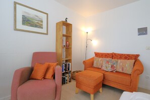 A living room with a red armchair, an orange sofa with floral cushions, a wooden bookshelf, and a floor lamp. - House Sylter Welle 5-39 WB (Sylt/Westerland)