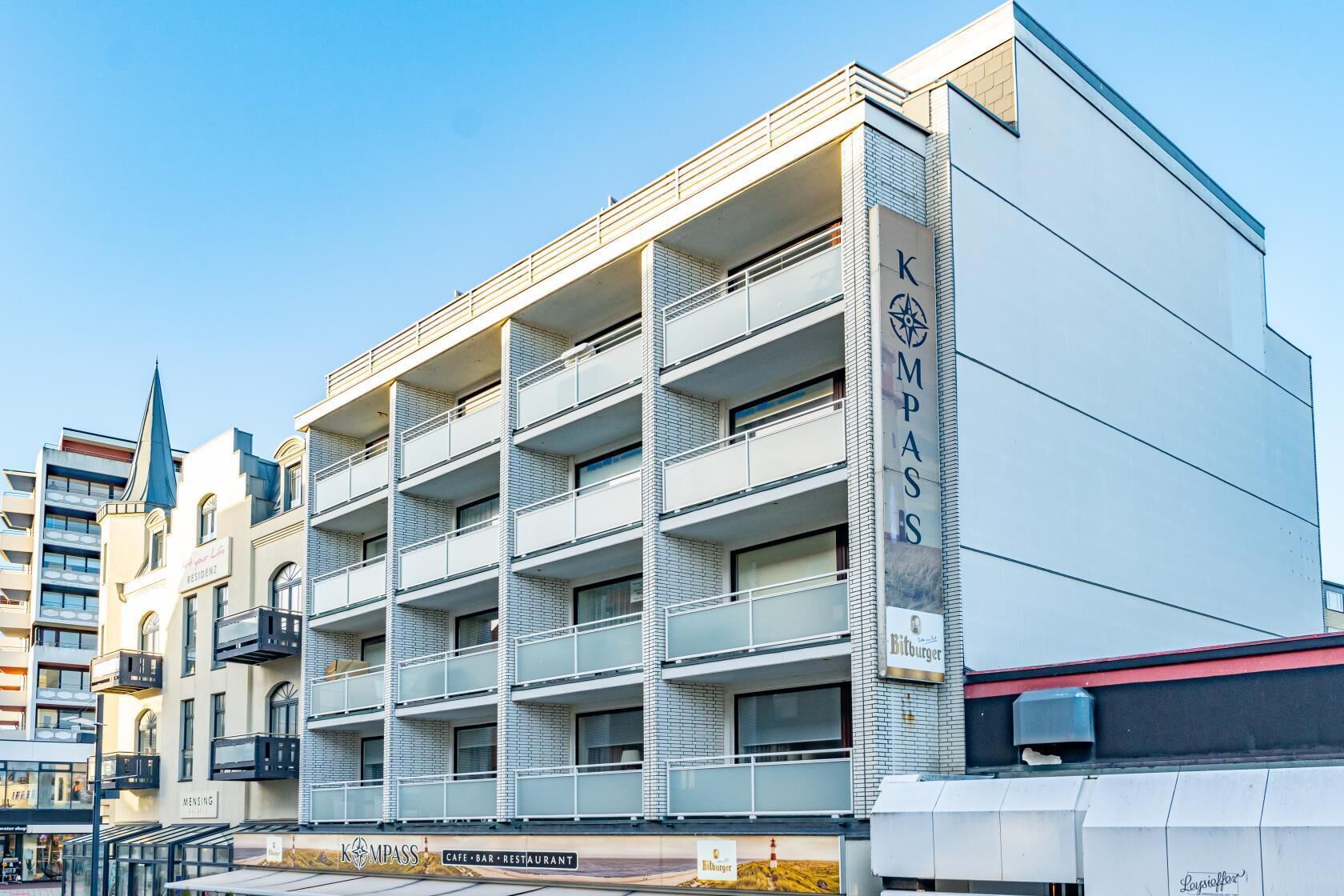Un edificio de varios pisos con un letrero que dice "KOMPASS" y "Café Bar Restaurante" en la planta baja.