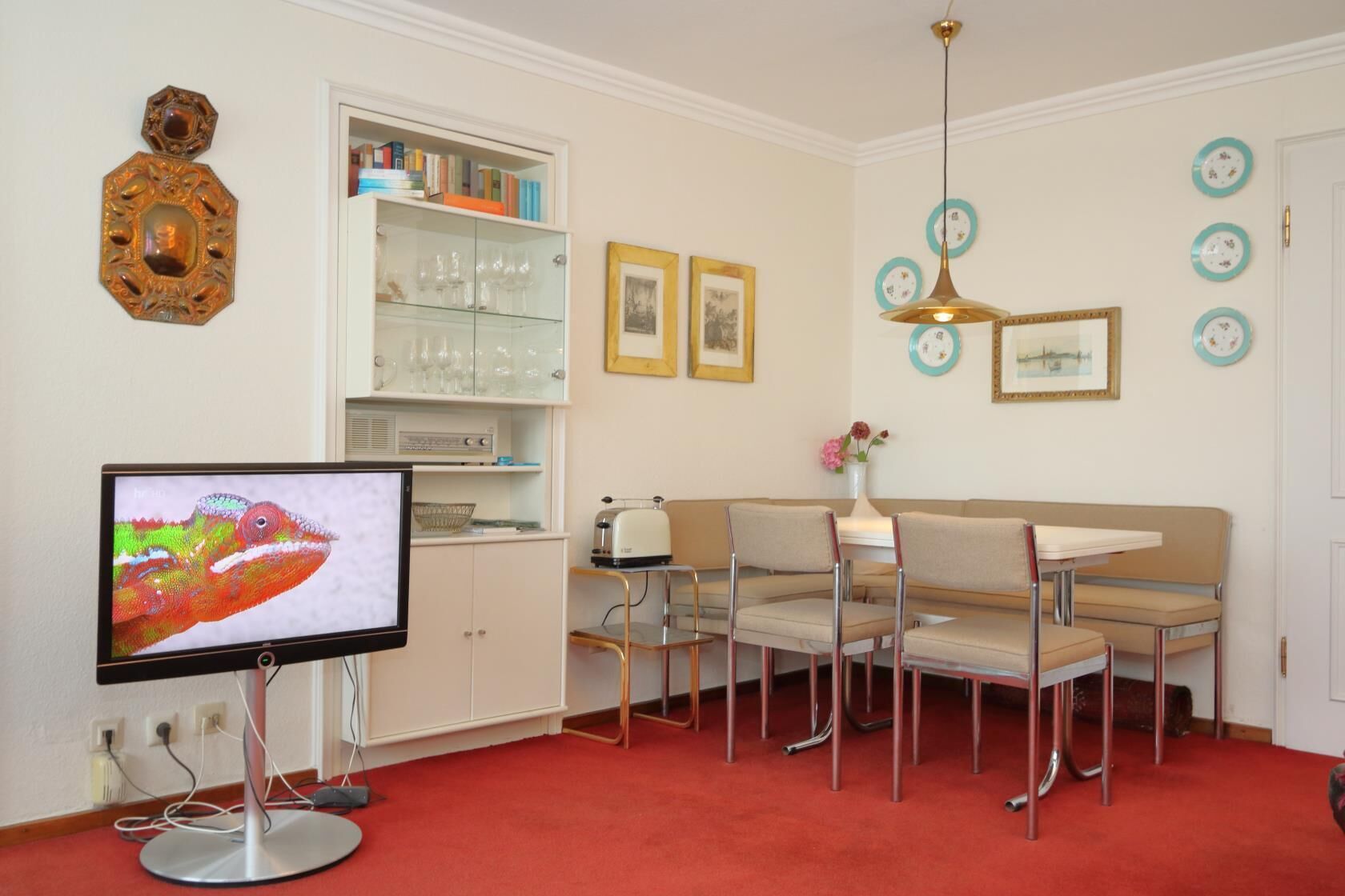 Quarto com televisão, armário branco com copos, área de refeições com cadeiras e tapete vermelho.