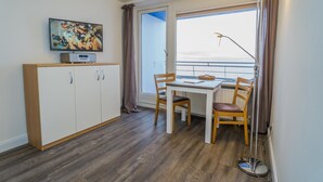Ett rum med trägolv, ett vitt bord med två stolar, en tv på ett skåp och ett stort fönster med gardiner.