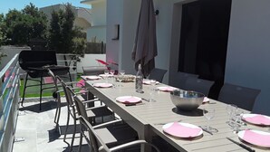 Zona de menjador a l'aire lliure amb una taula llarga ben preparada per a sis persones, envoltada de cadires, i una barbacoa i una ombrelles al fons.