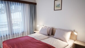 Een net opgemaakt bed met een rode deken, witte kussens en een houten hoofdeinde. Er staat een nachtkastje met een lamp en er hangt een ingelijste foto aan de muur.