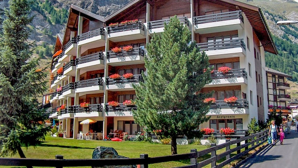 Een meerlaags hotel met bloembalkons, gelegen tegen een bergachtige achtergrond.