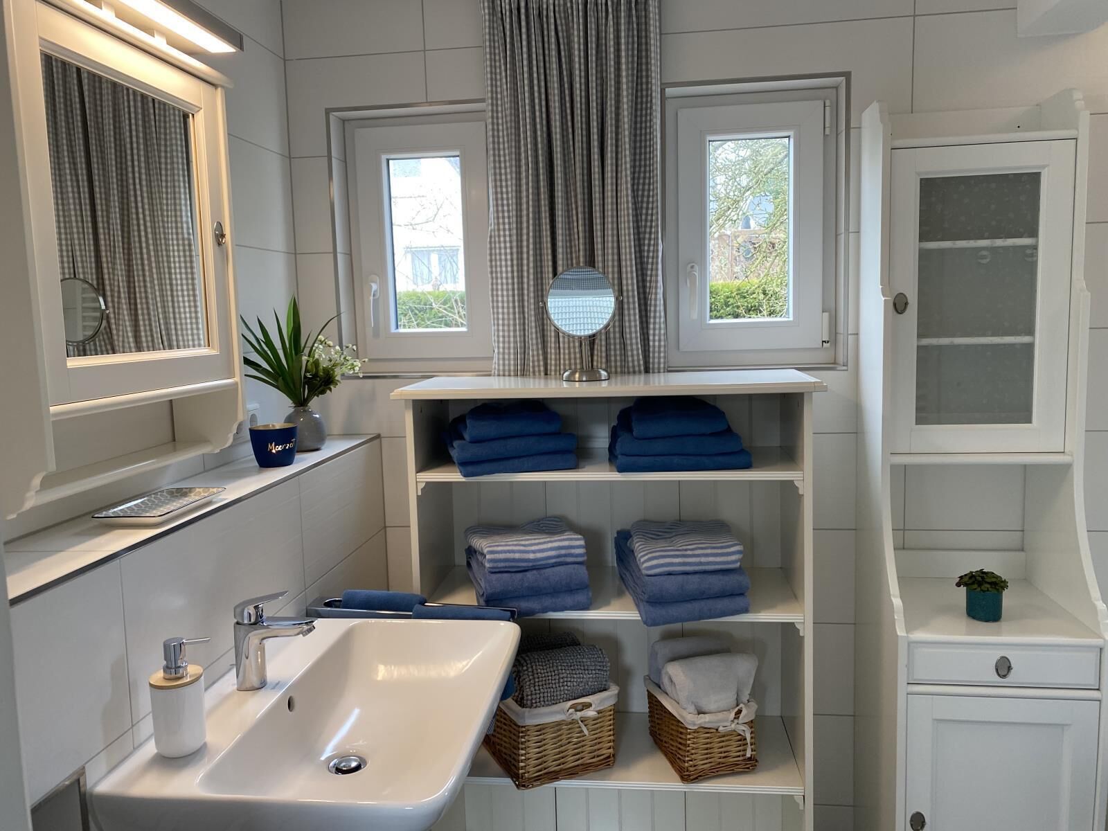 A bathroom with a white sink, mirror, and two windows with curtains. There are shelves with folded towels and baskets.