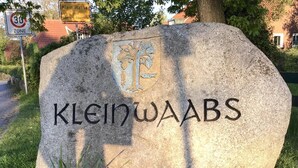 Un letrero de piedra con el nombre 'Klein Waabs' y un escudo, rodeado de hierba y flores.