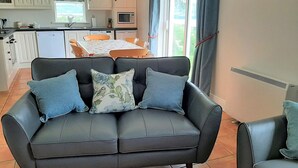 Een woonkamer met een zwarte leren bank, twee blauwe kussens, een eettafel met stoelen, een keuken met witte kasten, een magnetron en een raam met gordijnen.