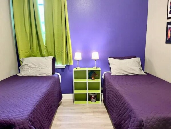 Una habitación con dos camas, cortinas verdes, una mesita de noche con dos lámparas y un estante verde.