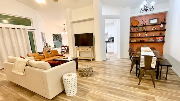 A modern living room with a sofa, coffee table, and ottoman. A dining area with a wooden table and chairs is visible. A TV is mounted on the wall. - The Sanford Ave House with fully-fenced yard (Sanford)