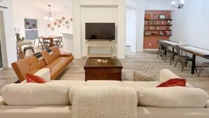 A modern living room with a sofa, coffee table, and TV. There is a dining area with chairs and a table. - The Sanford Ave House with fully-fenced yard (Sanford)