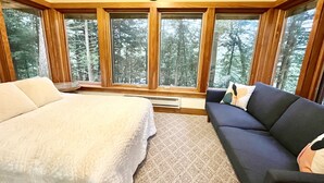 A bedroom with a large bed, a dark-colored sofa, and a view of a forest through large windows.
