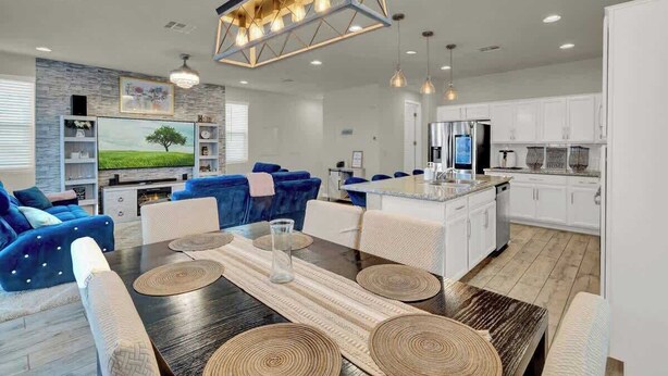 A modern living room with a dark wood dining table, blue sofas, and a kitchen area with white cabinets and a stainless steel refrigerator.