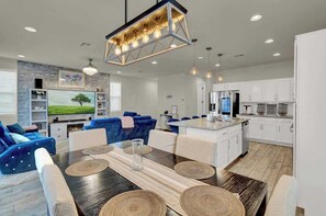 A modern living room with a dark wood dining table, blue sofas, and a kitchen area with white cabinets and a stainless steel refrigerator.