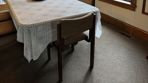 A dining area with a table covered by a lace tablecloth, wooden chairs, a chandelier, and a window with a view of greenery.