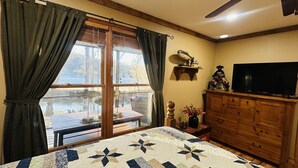 A room with a large window, a bed with a quilt, a wooden dresser, and a television. - Adorable, cozy, waterfront cabin on the River, fish, kayak, tube from property. (Mineral Bluff)