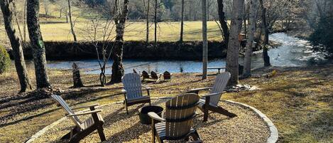 Un espace de détente circulaire, composé de fauteuils Adirondack et d’un foyer, entouré d’un chemin de gravier et d’arbres, et offrant une vue sur une rivière.