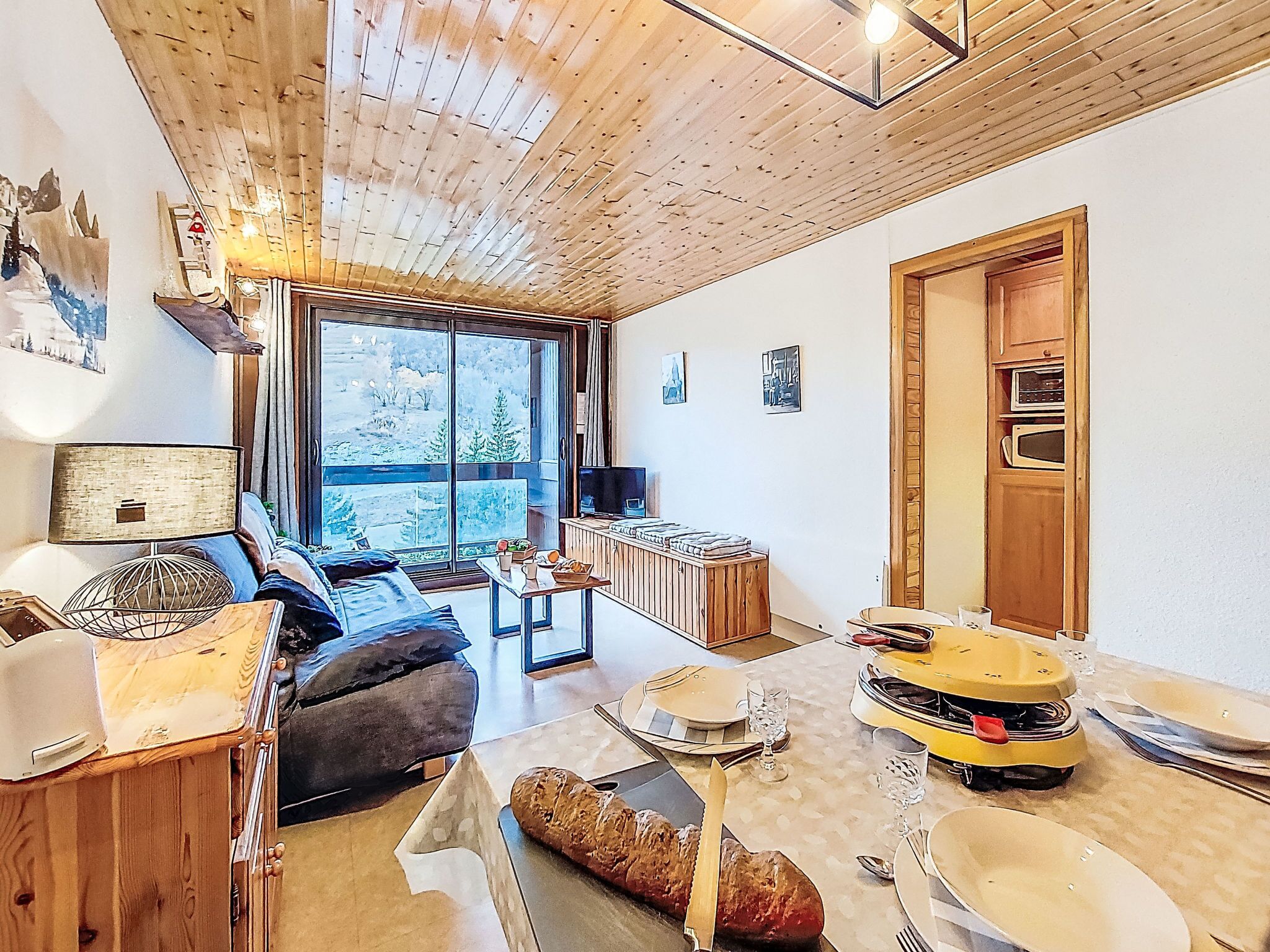 A cozy room with a wooden ceiling, a large window with a view of trees, a dining table set with plates, glasses, and a loaf of bread.