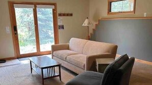 A living room with a sofa, a chair, a coffee table, and a lamp. There is a window with a view of trees and a door leading outside.