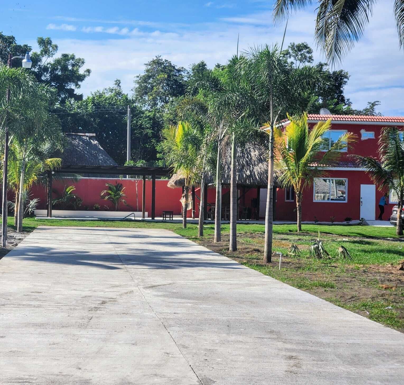 A pathway leading to a red building with a tiled roof, surrounded by palm trees and greenery.
