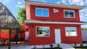 A two-story red house with a white door and windows, a thatched roof structure to the left, and a small garden area in front. - VILLA CLAUDIAS (Taxisco)