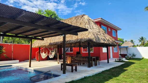A pool area with a thatched-roof structure, a red building, and a clear blue sky. - VILLA CLAUDIAS (Taxisco)