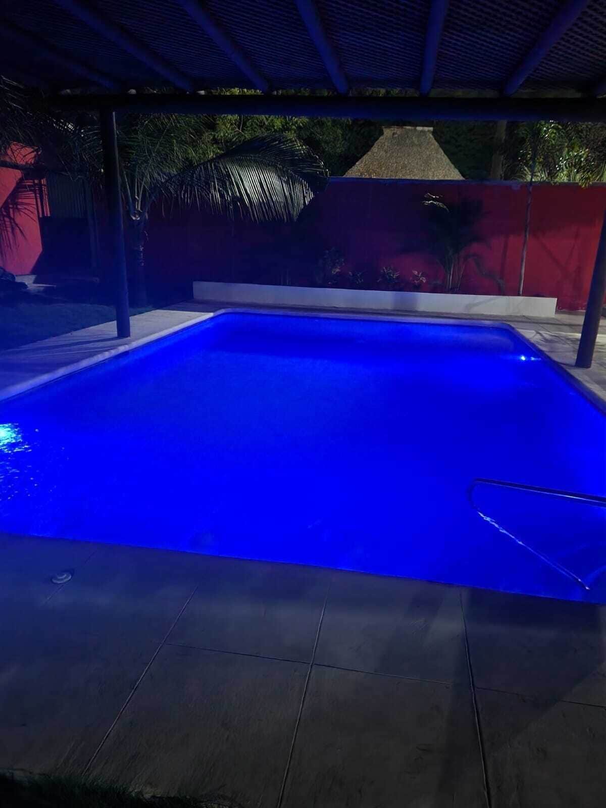 A swimming pool with a blue-lit interior, surrounded by a covered area with a red wall and some greenery in the background.