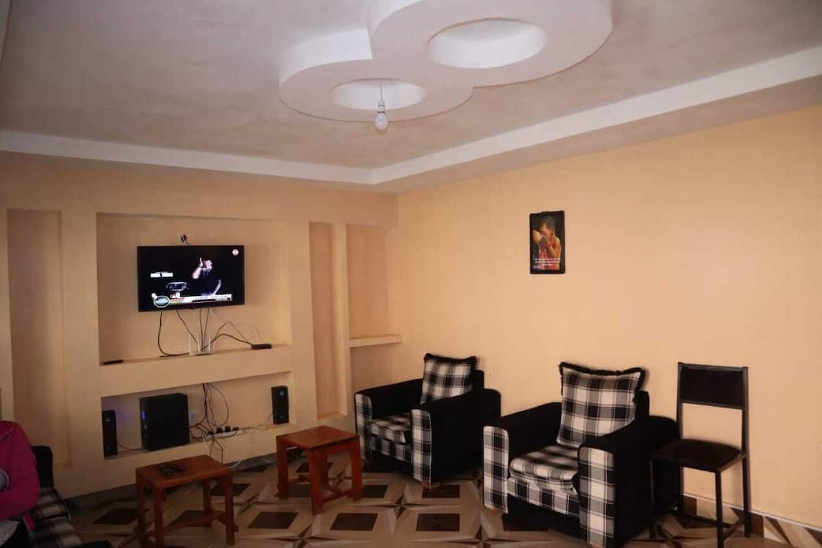 A living room with a flat-screen TV mounted on the wall, two plaid-covered armchairs, a wooden coffee table, and a black floor lamp.