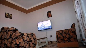 Un salon avec un téléviseur fixé au mur, deux fauteuils à motifs, une petite table blanche et un rideau à motifs.