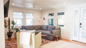 A modern living room with a sectional sofa, a small table, a potted plant, and a large window. - Irie on Erie B (Folly Beach)
