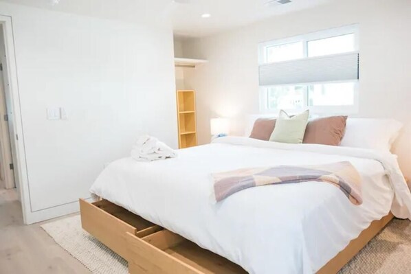 Ein Schlafzimmer mit einem Bett, einer Schublade aus Holz darunter, einem Fenster mit Jalousien und einer weißen Wand.