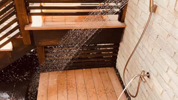 Douchegedeelte met een houten bankje, een metalen douchekop en een witte baksteenmuur.