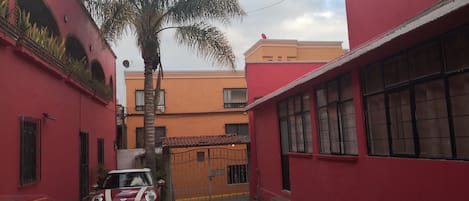 A narrow alleyway with a red building on the left and a yellow building on the right. A palm tree is in the center, and a red car is parked on the left side.
