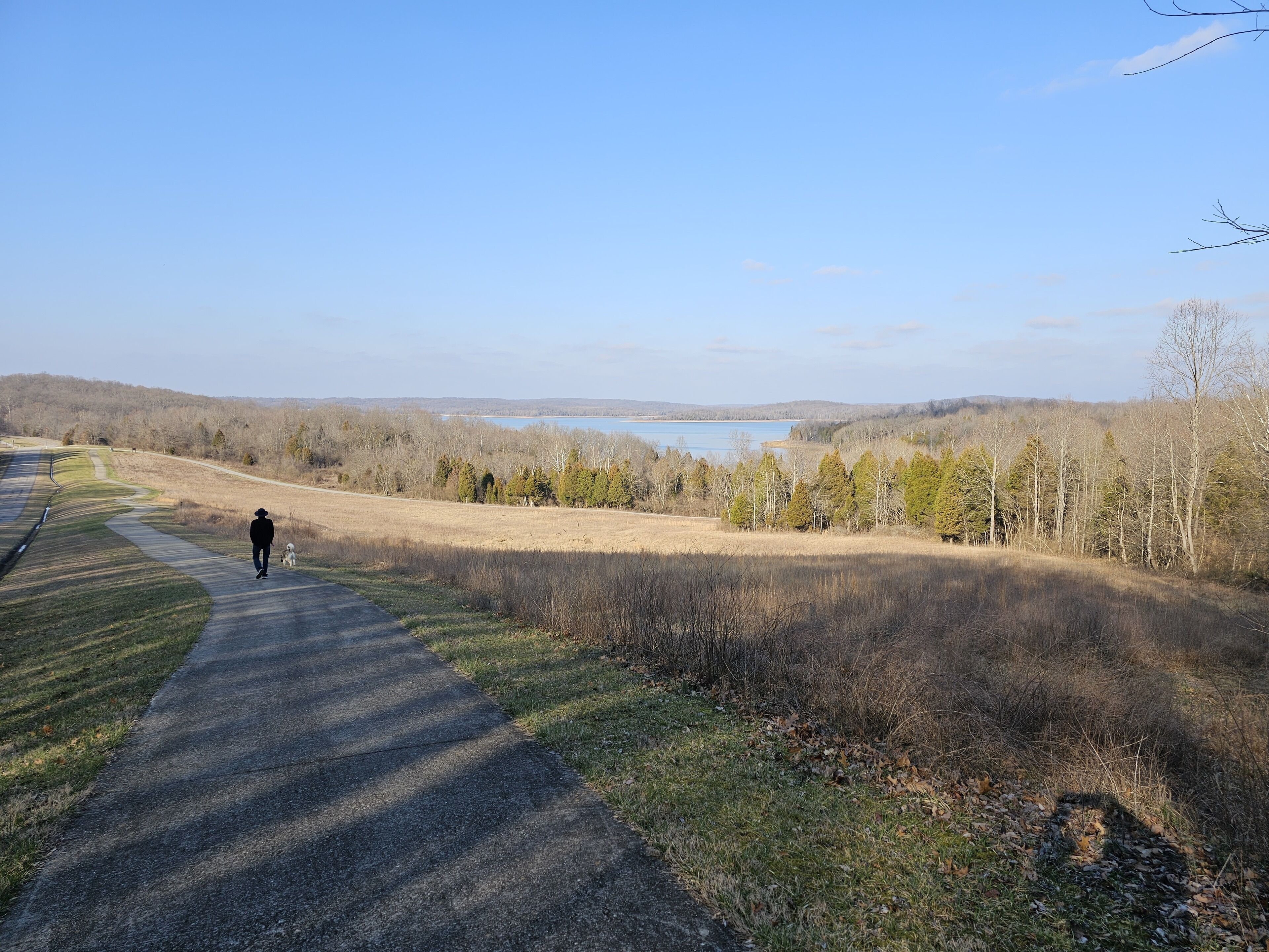 A paved path leading to a body of water with trees and hills in the background.
