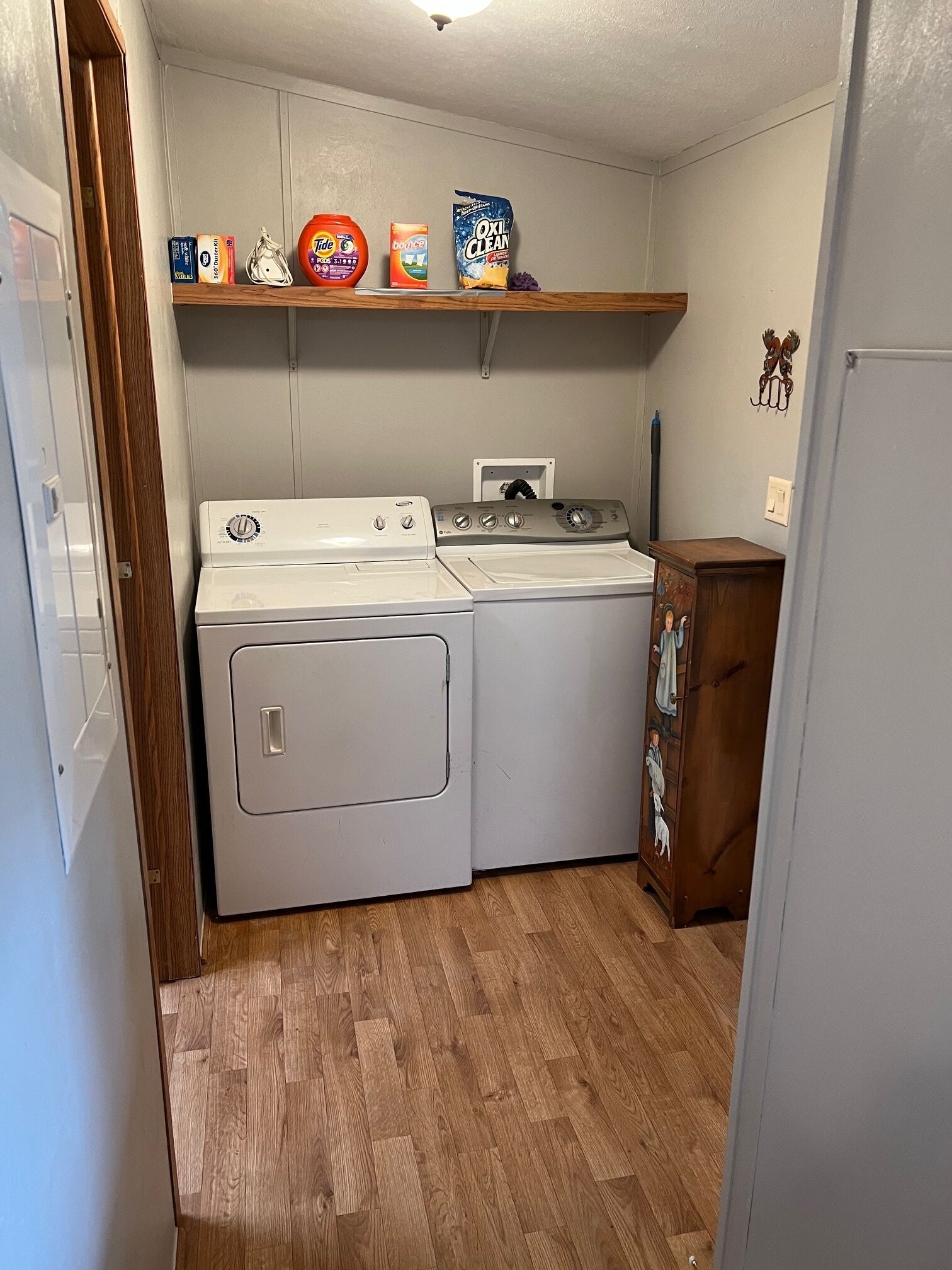 A laundry room with two washers and dryers, a shelf with laundry supplies, and a wooden cabinet with a religious painting.