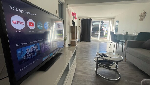 A living room with a large flat-screen TV displaying streaming services. There is a coffee table with a remote control, a sofa, and a dining area in the background. - Apparemment à Côté du Meet (Cornebarrieu)