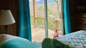 Una stanza con un letto, una scrivania in legno, una lampada e vista su una terrazza e montagne attraverso una porta di vetro.