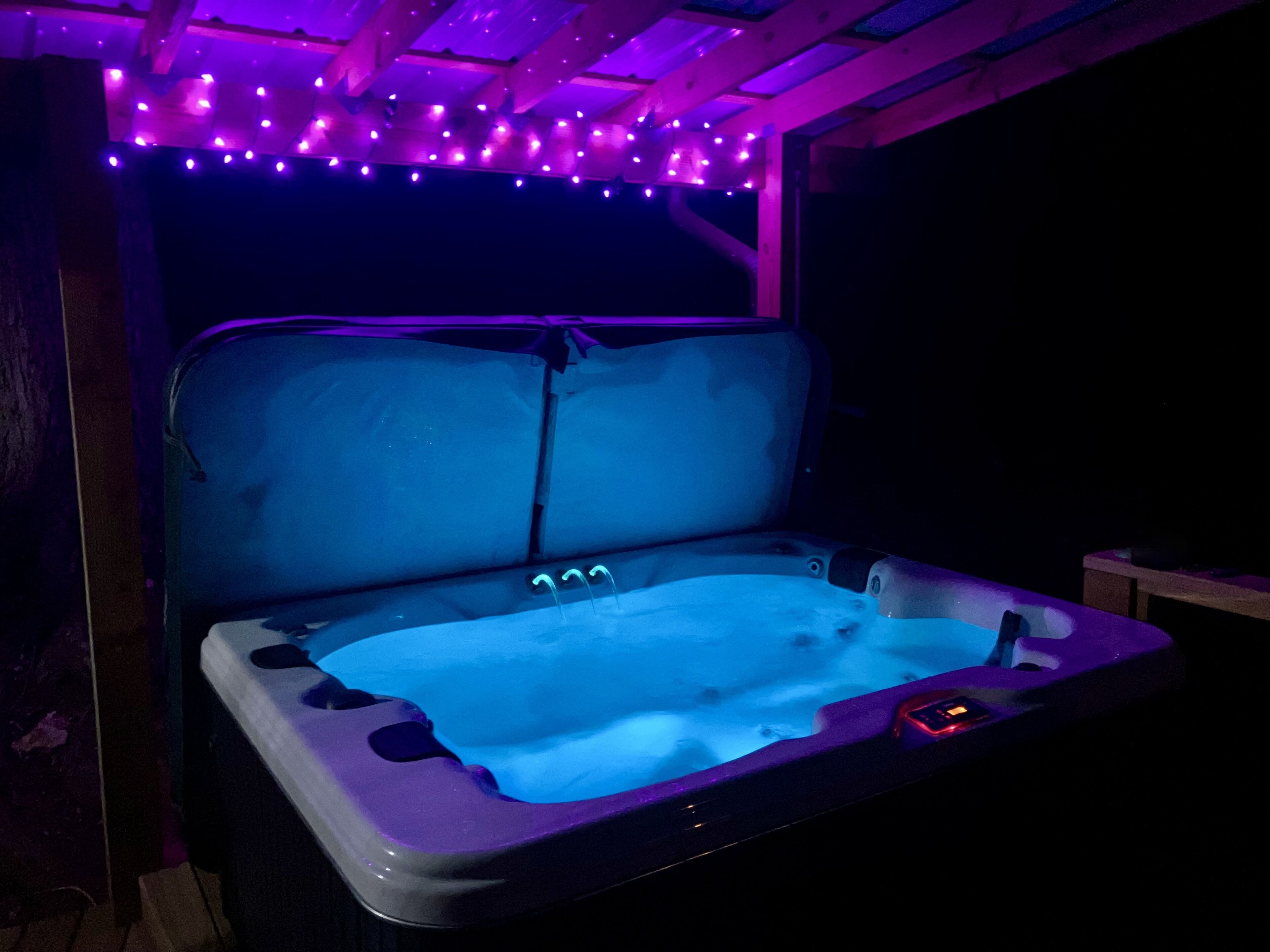 Un jacuzzi con interior azul, rodeado de vigas de madera e iluminación púrpura.