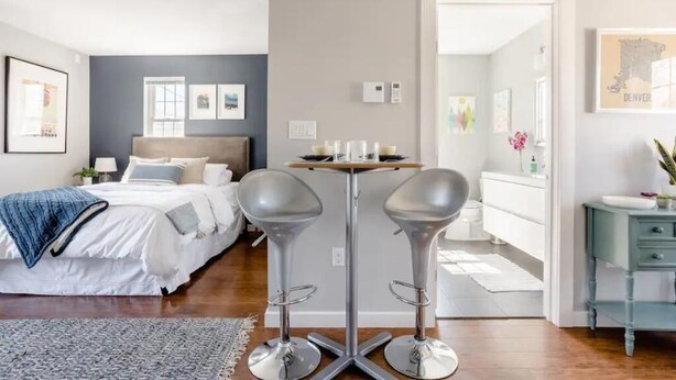 Une chambre d’hôtel moderne avec un lit, une petite table avec des bougies, deux tabourets de bar et une salle de bain visible à travers une porte ouverte.