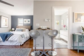 Une chambre d’hôtel moderne avec un lit, une petite table avec des bougies, deux tabourets de bar et une salle de bain visible à travers une porte ouverte.