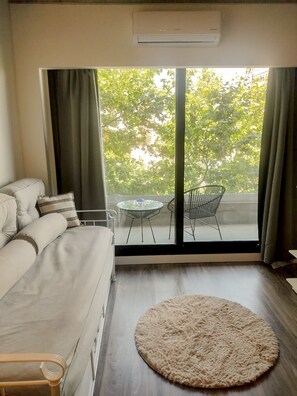 A modern living room with a sofa, a round rug, a glass table, and a view of trees through large windows.