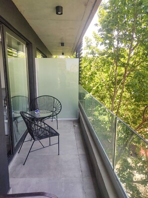 Un balcón con barandilla de vidrio, una mesa con dos sillas y vistas a árboles.