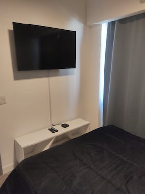 Un dormitorio con cama, televisor montado en la pared, mesita de noche con regleta de enchufes y ventana con cortinas.