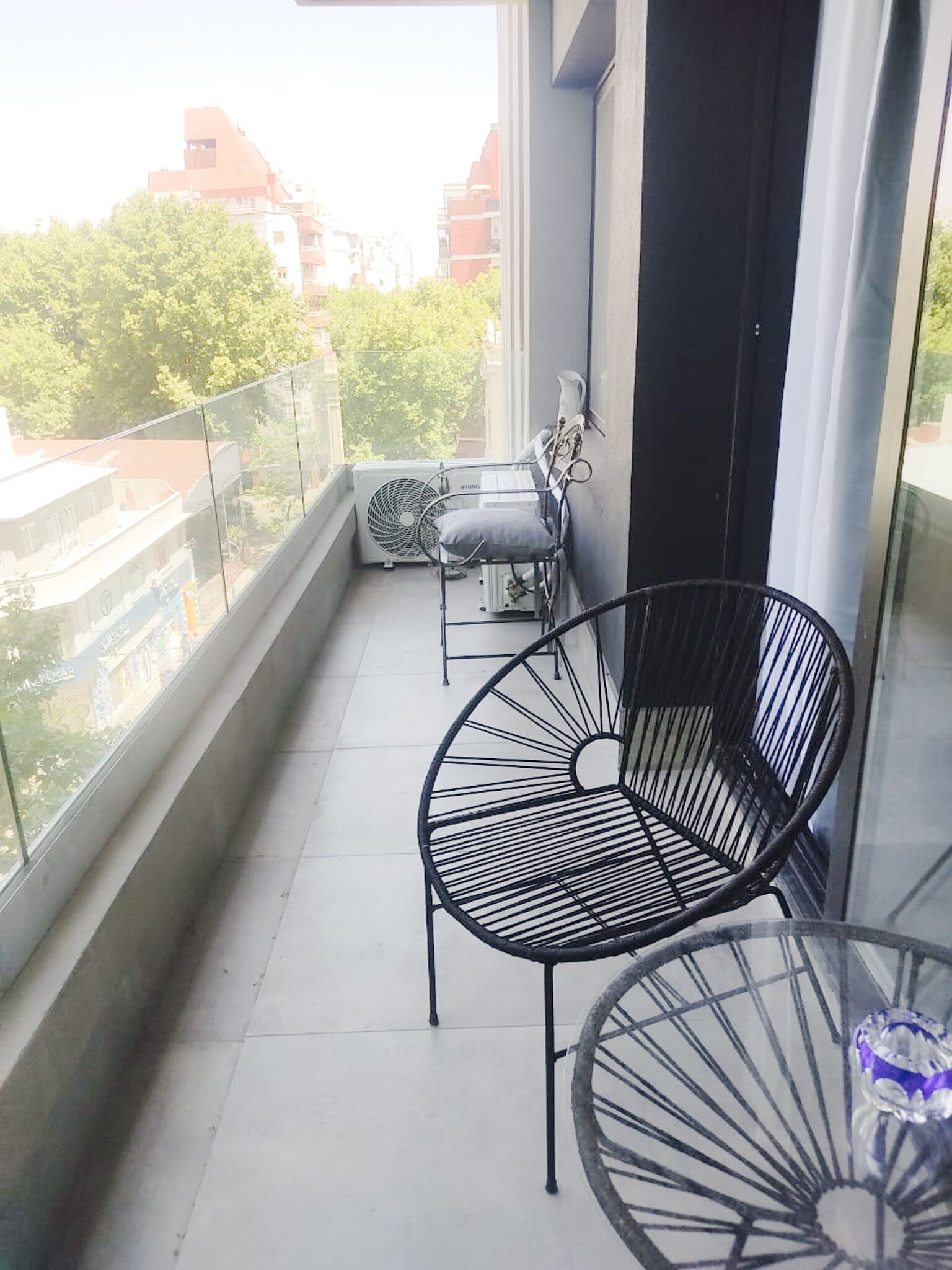 Een balkon met een glazen hekwerk, twee zwarte metalen stoelen, een klein tafeltje en een witte ventilator.