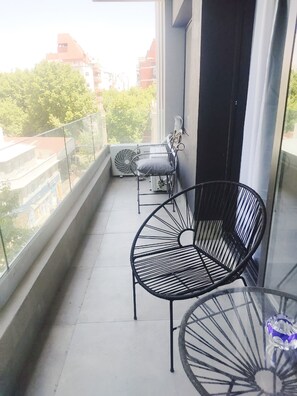 Uma varanda com guarda-corpo de vidro, duas cadeiras de metal preto, uma mesa pequena e um ventilador branco.